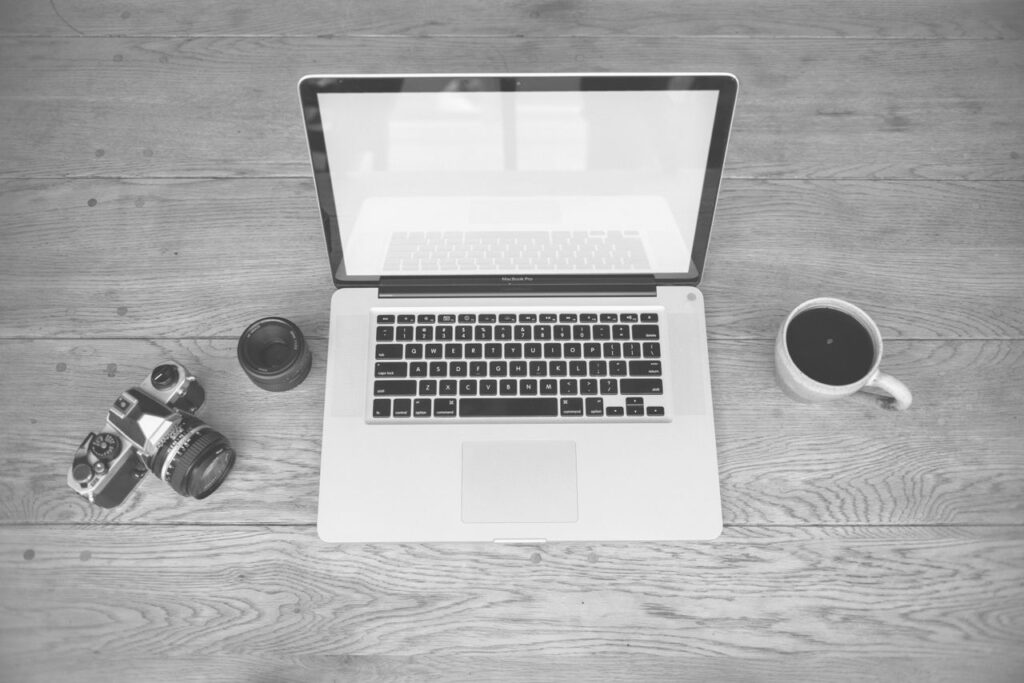 Monochrome image of a laptop, camera, lens, and coffee cup on a wooden desk